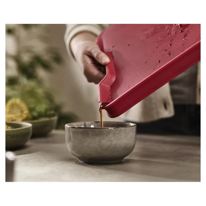 Person using a red plastic chopping board to pour liquid into a grey bowl in kitchen setting