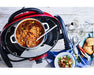 Portable outdoor grill with brown trivet kit, stew pot, bread, and serving utensils on table.